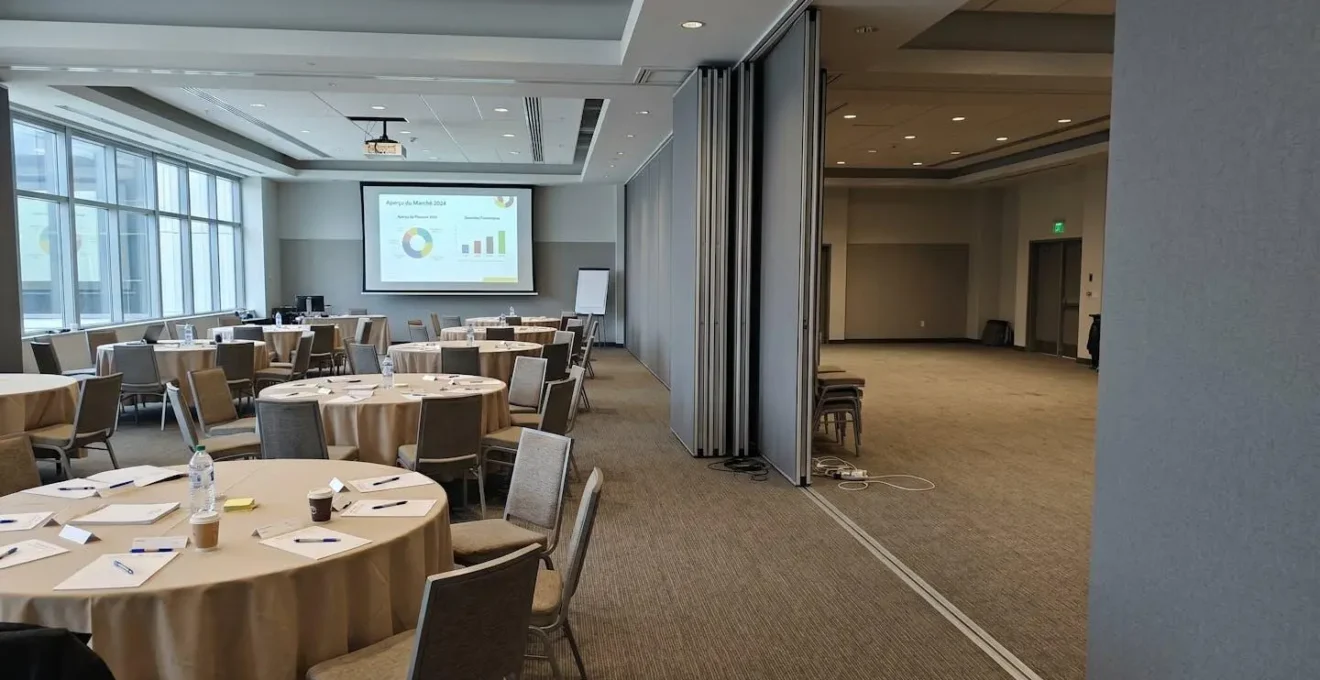 Salle de conférence contemporaine vide avec rangées de tables rondes et chaises noires, baignée de lumière naturelle par de larges fenêtres